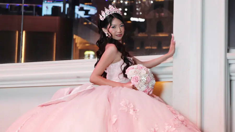 Quinceanera portrait in a pink gown beside a bright window.